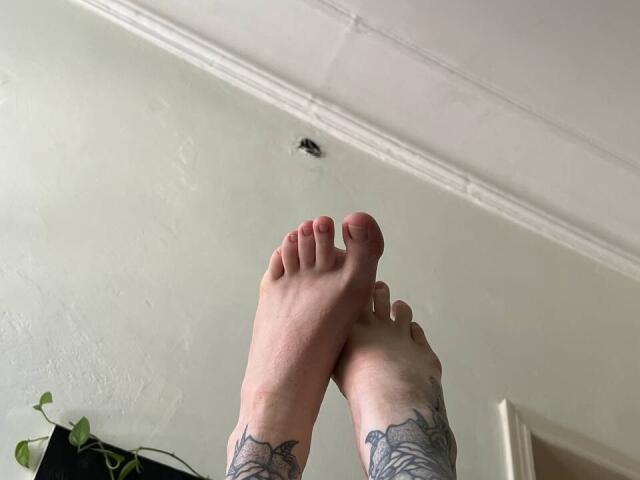 Mettis' Füße, mit Tattoos verziert, waren an den Knöcheln verschränkt, wobei die nackten Fußsohlen und Zehen zu sehen waren. Über ihr befand sich eine kleine Öffnung in der Decke. Mettis hatte Tattoos an den Waden..