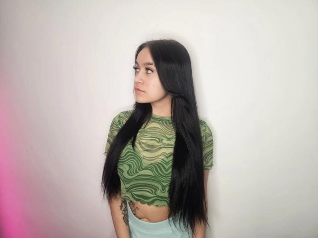 Aisha, con lunghi capelli neri, indossa un top corto verde con motivi ondulati e pantaloni azzurri, è in piedi contro un muro bianco, guarda di lato e ha un tatuaggio sul braccio destro..