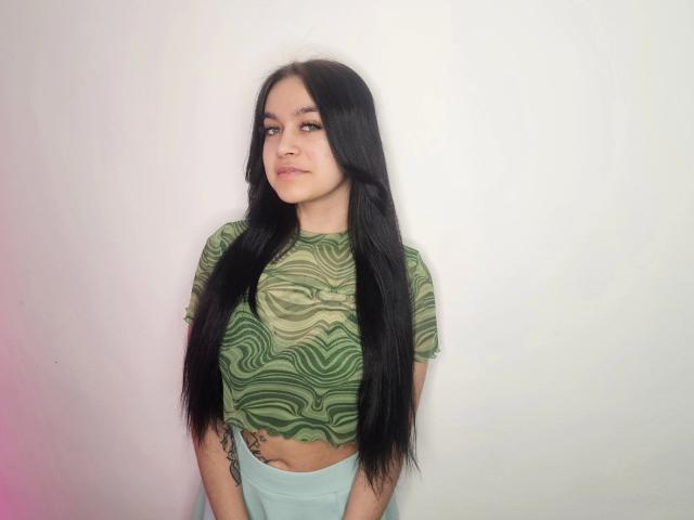 Aisha, met lang zwart haar, een groene crop top met zebrapatroon en lichtblauwe broeken met hoge taille, toont een tatoeage op haar rechterheup, en staat tegen een effen witte muur..