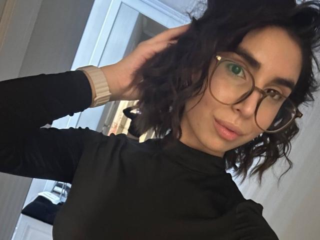 TameikaMacclellan with curly black hair, green eyes, and glasses, wearing a black turtleneck, hand in hair, smiling, upper body, soft light, subtle makeup, sexy, attractive.