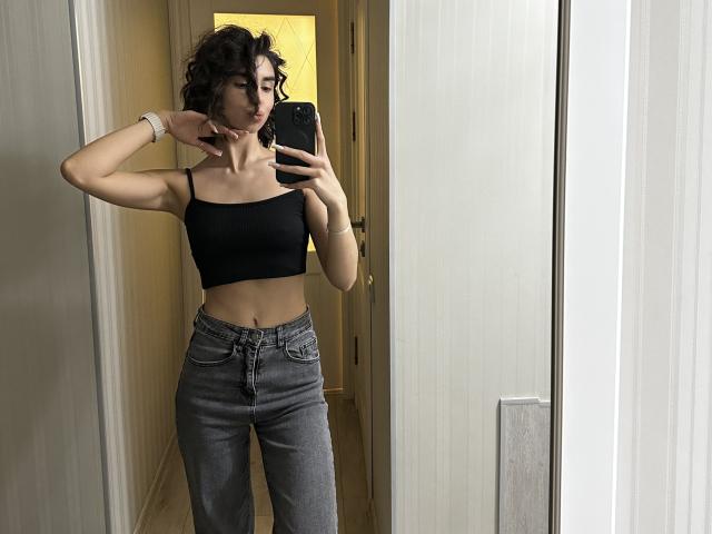 TameikaMacclellan, mirror selfie, wearing black crop top, showing small tits, gray high-waisted jeans, wavy dark hair, white wristband, holding phone, hallway, wooden floor, serious expression.