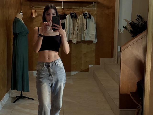 TameikaMacclellan with medium tits in a black crop top, holding phone, wearing light blue jeans, standing in a wooden-walled room, mirror selfie, green dress on mannequin.