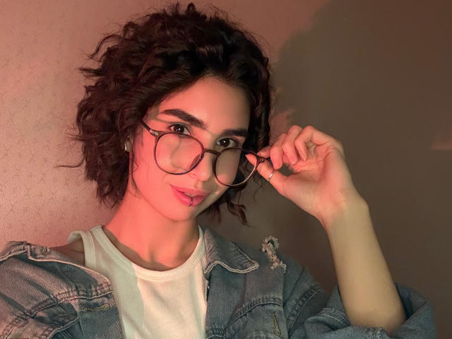 TameikaMacclellan with curly dark brown hair, wearing glasses, a denim jacket, and white shirt, adjusting her glasses with a slightly pouty expression, under pinkish lighting.