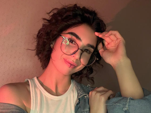 TameikaMacclellan with curly brown hair, wearing glasses, a white tank top, and a blue denim jacket, smiling and touching her hair, illuminated by red and yellow light.