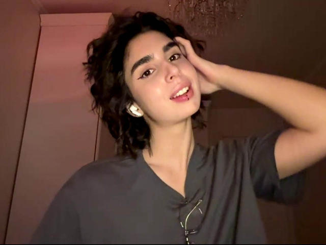 TameikaMacclellan with curly dark hair, wearing a grey shirt, smiling with hand on head, pinkish-red light, visible glasses on shirt, light skin, slightly parted lips, looking at camera.