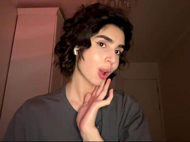 TameikaMacclellan with curly black hair, wearing a gray shirt, touching her lips with her hand, has white earbuds, and a slightly open mouth, looking at the camera.
