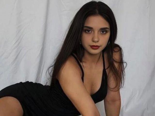 ScarlettX, une femme séduisante avec de longs cheveux noirs, portant une robe noire, penchée en avant, montrant sa poitrine, avec une expression sérieuse, du rouge à lèvres et des yeux marron, sur un fond blanc..