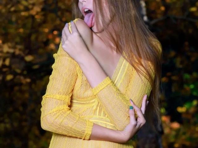 ScarlettX, aantrekkelijk met lang bruin haar, gele kantjurk, tong uit, bijt in haar eigen schouder, hand met blauwe nagel, gele ring, herfstbos op de achtergrond, laat een ontbloot schouder zien..