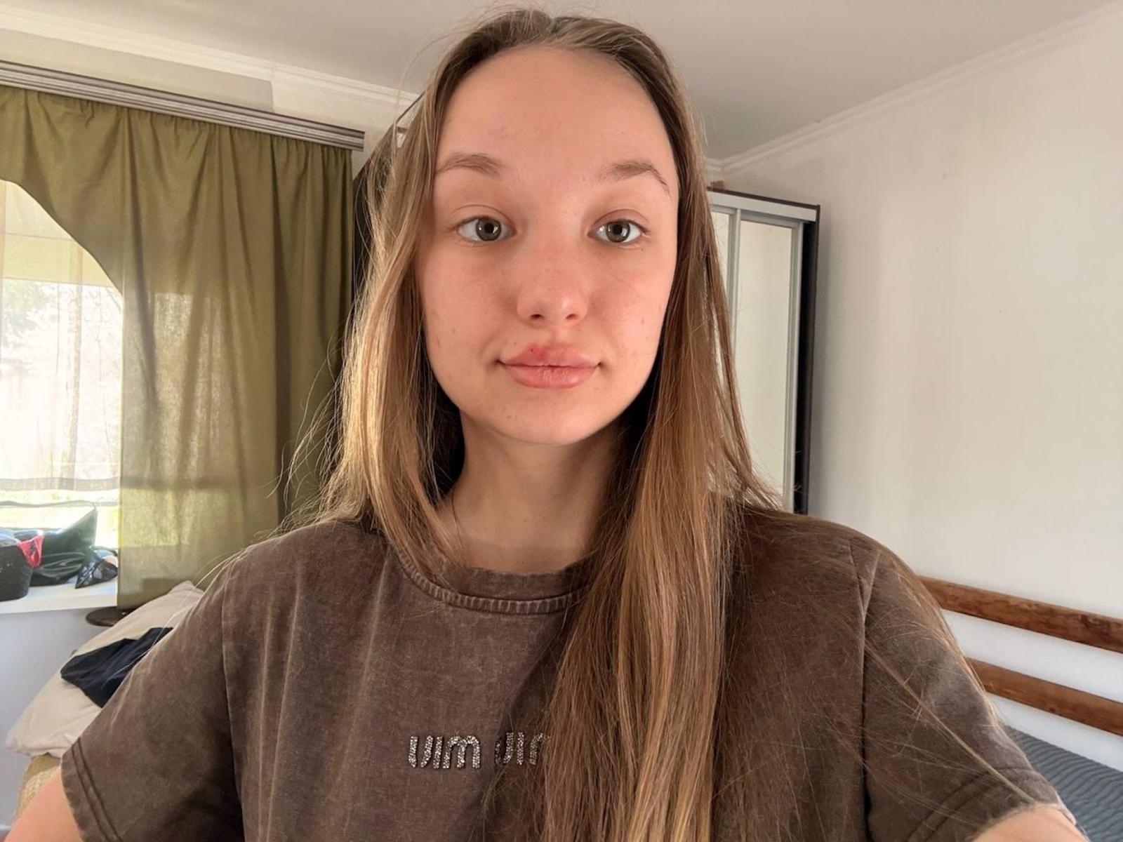 RandiMatute has long brown hair, green eyes, and a slightly pouty face wearing a brown shirt. She's standing in a bright room with green curtains.