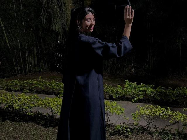 MiluSky, with long dark hair in a ponytail, wearing a black dress, holds a black hat up, smiling, in a dark, forested area with a lit path.