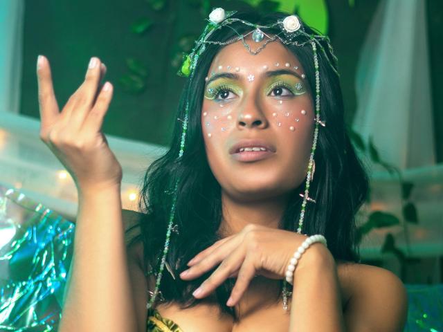 RoxanneHan with green eyeshadow, white face dots, and a jeweled headpiece, raising her right hand, showing off her tits, wearing a bead bracelet, and looking up.