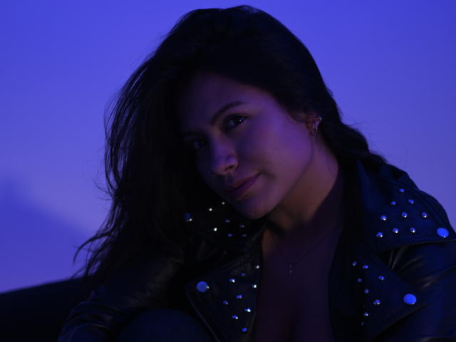 Laura Miller, met lang donker haar, draagt een zwarte leren jas met studs, kijkt naar beneden met een lichte glimlach, en wordt verlicht door blauw licht, waardoor haar schattige neus en kleine oorbellen zichtbaar zijn..