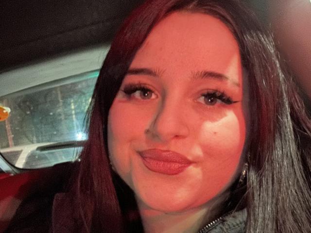 LucieRed has red lipstick, long eyelashes, and a slight smile, with dark brown hair and fair skin. She's in a car, illuminated by red light.