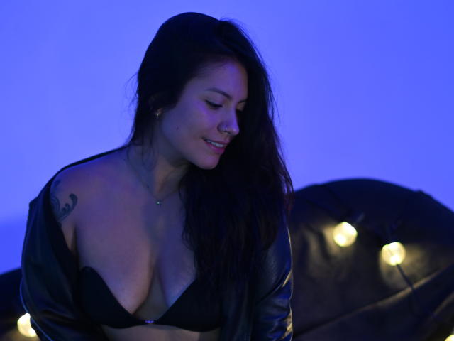 Laura Miller, con i capelli scuri, indossa un reggiseno nero e una giacca di pelle aperta, mostrando il seno, sorride guardando verso il basso, illuminata di blu, con luci a stringa e una poltrona di pelle sullo sfondo..