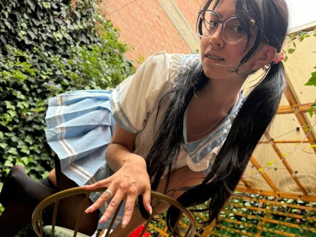 VioletXBloom with long black pigtails, wearing glasses, a blue and white schoolgirl outfit, and black tights, leaning on a chair with a slight smile, showing her tits.