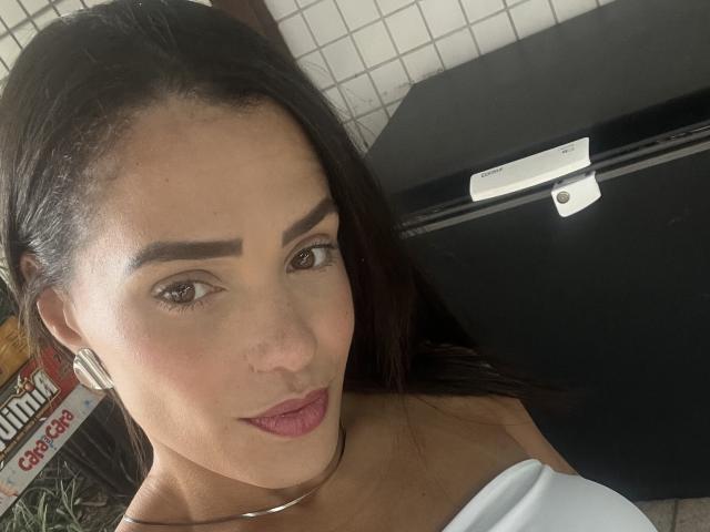 StellinhaSky, con cabello oscuro, cejas pobladas y lápiz labial rosa, mostrando sus senos en una blusa blanca con un hombro descubierto, está de pie junto a una maleta negra..
