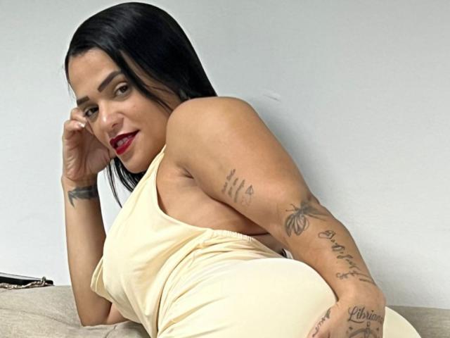 StellinhaSky mit schwarzen Haaren, rotem Lippenstift und Tätowierungen, trägt ein gelbes Kleid, liegt auf einer Couch, schaut mit einem leichten Lächeln in die Kamera und zeigt ihr Gesäß..