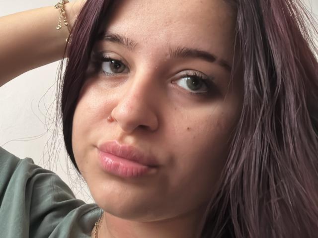 LucieRed's face, showing light green eyes, pink lips, and light skin with a few moles. She has dark purple hair and a gold bracelet on her right wrist.