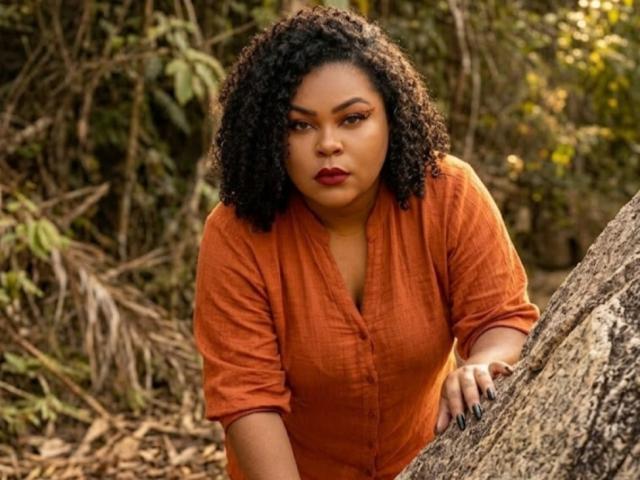 MariaLuissa69, avec ses cheveux noirs bouclés, ses lèvres rouges pulpeuses et sa peau brun clair, porte une chemise orange à boutons, est appuyée contre un arbre dans une forêt et a l'air sérieuse..