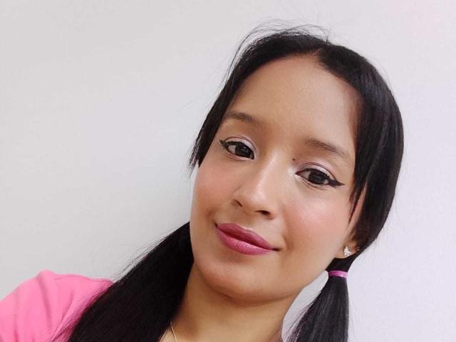 Shanel White ha la pelle marrone chiaro, due trecce nere e rossetto rosa con eyeliner ad ali di farfalla, indossa una camicia rosa e sorride dolcemente alla fotocamera. Ha degli orecchini piccoli e rotondi..