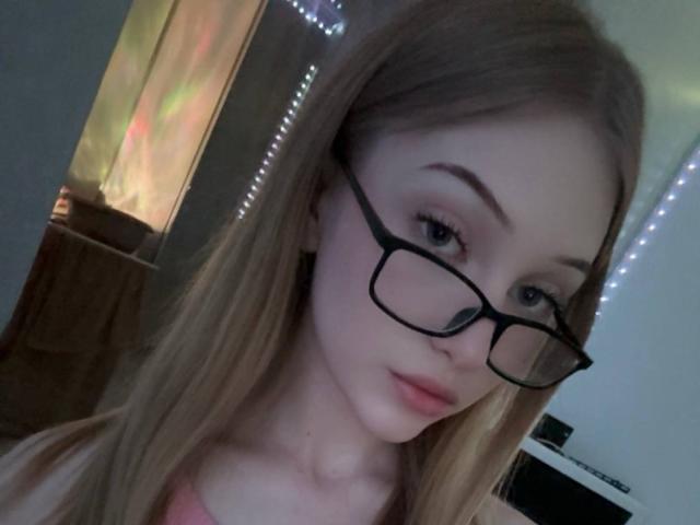 Carlyn Freiser, con lunghi capelli biondi, indossa occhiali neri, ha la pelle chiara, rossetto rosa e una maglietta rosa, guarda la fotocamera con un leggero sorriso, in una stanza con una luce soffusa..