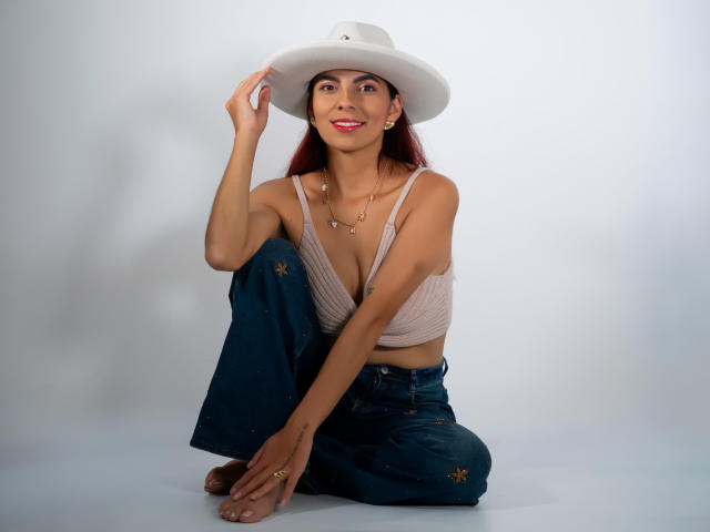 CarlaTorres, sentada, usando um chapéu branco, cropped rosa, mostrando seios pequenos, calça jeans com padrões dourados, sorrindo, batom vermelho, pele morena, cabelo longo e ruivo, descalça, mão no chapéu..