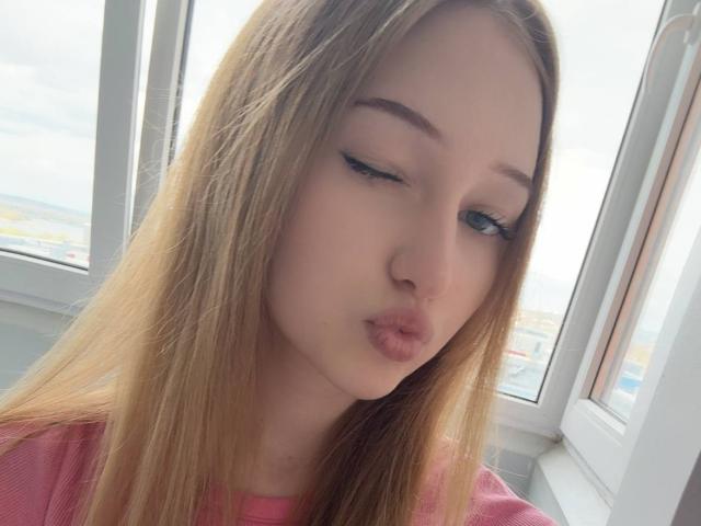 CarlynFreiser with pouty lips, blonde hair, blue eyes, and pink shirt, winking with subtle eyeliner, soft pink skin, and a cute, slightly awkward expression.