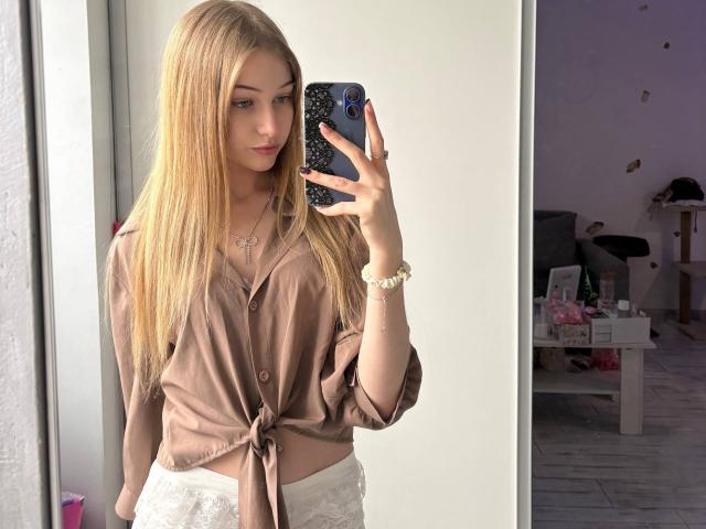 Carlyn Freiser, con lunghi capelli biondi, una camicia marrone con bottoni, pantaloni bianchi in pizzo, che tiene in mano un telefono, un selfie allo specchio, con un leggero sbuffo, una custodia per telefono blu, un braccialetto bianco, in una stanza ben illuminata..