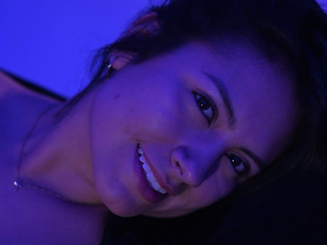 Laura Miller hat ein süßes Lächeln, dunkelbraunes Haar und eine kleine Sommersprosse auf ihrer rechten Wange. Sie schaut mit blau getöntem Licht in die Kamera..