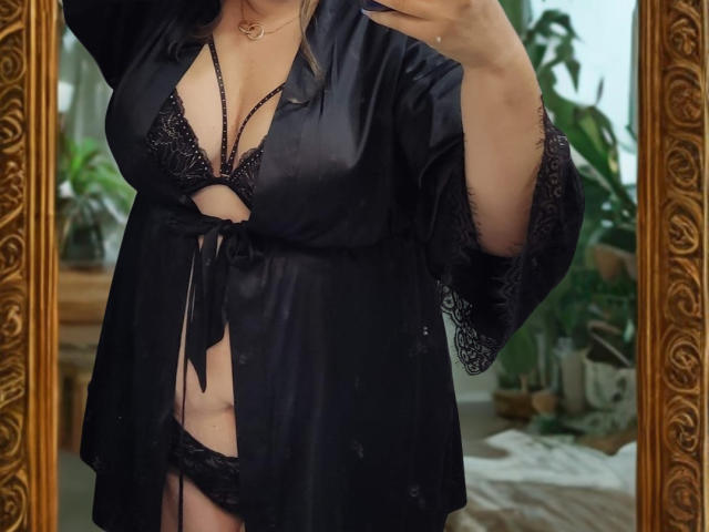 NettyElvie trägt einen schwarzen Spitzen-BH und passende Unterwäsche, dazu einen seidigen schwarzen Bademantel. Auf einem Selfie im Spiegel sind ihre Brüste zu sehen und ein kleiner Blick auf ihre Vulva ist erkennbar..
