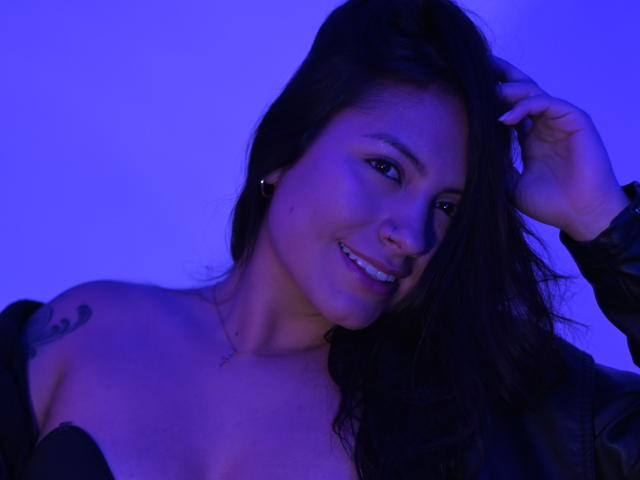 LauraaMillers with dark hair, smiling, blue-lit, wearing black leather, hand in hair, visible tattoo on right shoulder, cute, pretty, tits slightly visible, casual, confident.