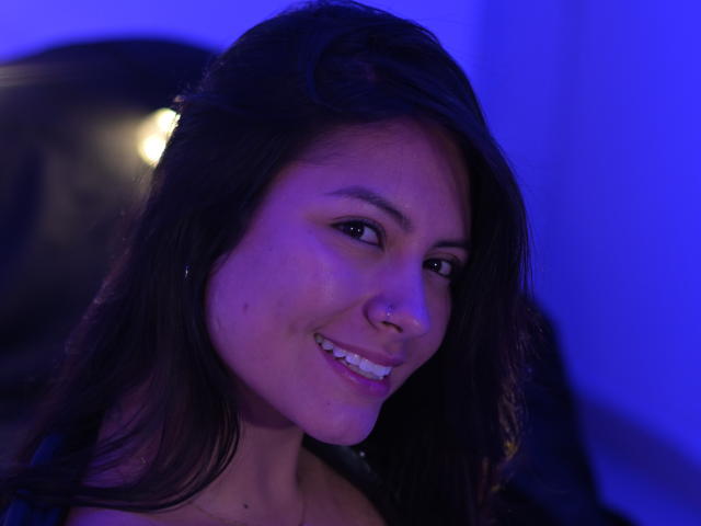 Laura Miller, con cabello castaño oscuro, sonriendo, con un piercing en la nariz y maquillaje discreto, bajo una iluminación azul, mostrando sus dientes blancos y pequeños, y una parte de sus glúteos ligeramente visible en el fondo..