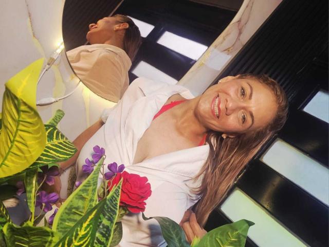 MirandaCeleste met blond, nat haar, lachend, gekleed in een witte gewaad, waarbij een roze bh zichtbaar is, staand in de buurt van groene planten en paarse bloemen, weergegeven in een spiegel..