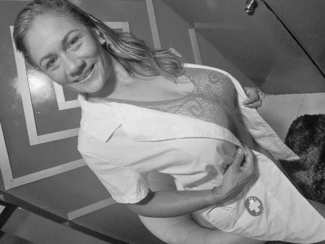 MirandaCeleste, avec des cheveux ondulés, souriante, portant un soutien-gorge en dentelle et une blouse d'hôpital, exposant ses seins avec une main, l'autre tenant la blouse. Photo en noir et blanc..