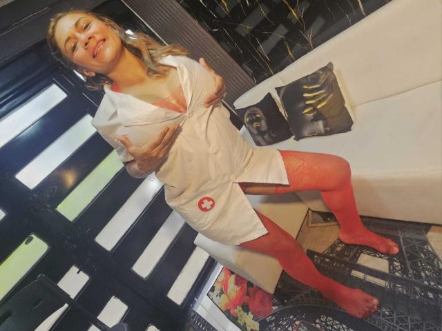 MirandaCeleste in een witte verpleegsterjurk, rode netpanty's en een rode kanten beha, die haar borsten vasthoudt, staand in een moderne kamer, met een glimlach en haar tong uitgestoken..