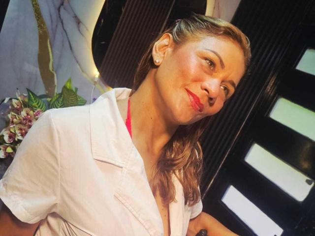 MirandaCeleste, con i capelli castano chiaro, rossetto rosso, camicia bianca e un leggero sorriso, che le conferisce un'espressione maliziosa, indossa piccoli orecchini, in un ambiente interno con una calda illuminazione..