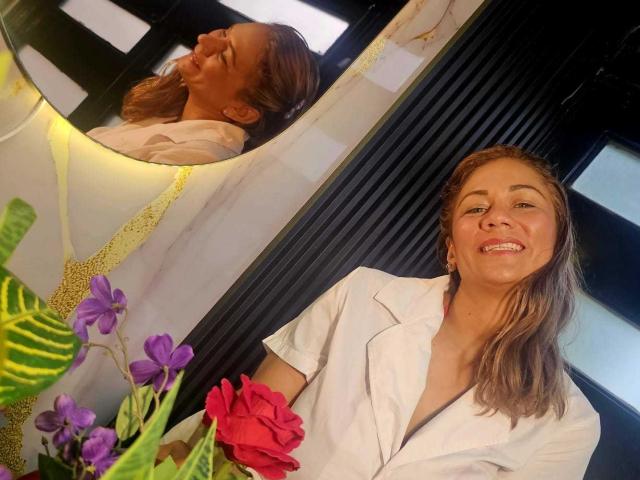 Miranda Celeste, mit einem Lächeln, braunes Haar, weißes Hemd, hält Blumen mit lila und rosa Blüten, ihr Profil wird in einem Spiegel widergespiegelt, in einem hellen Raum..