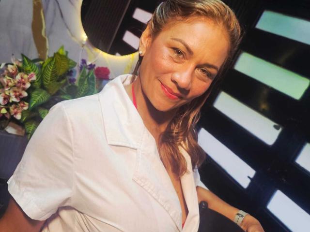 MirandaCeleste, con cabello rubio, sonriendo, vestida con una camisa blanca, mostrando ligeramente sus senos, con lápiz labial rojo, aretes y un reloj de pulsera, en un entorno con buena iluminación y fondo oscuro..