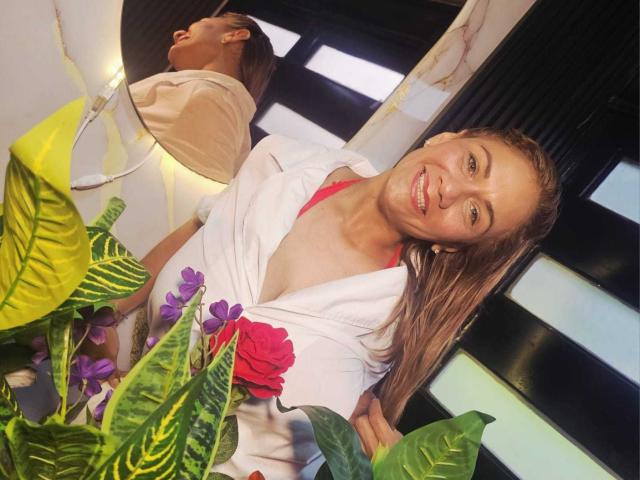 MirandaCeleste, avec ses cheveux blonds mouillés, souriante, portant une robe blanche avec un motif floral rouge, montrant sa poitrine, debout près de plantes vertes et violettes, reflet dans un miroir en arrière-plan..