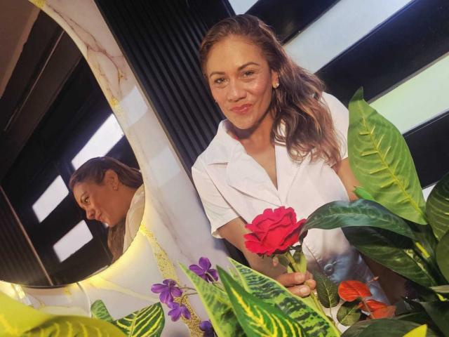 MirandaCeleste met golvend bruin haar, een wit shirt, die een rode roos vasthoudt, staat in de buurt van groene bladeren en paarse bloemen, glimlacht lichtjes en wordt weerspiegeld in een spiegel aan de linkerkant..