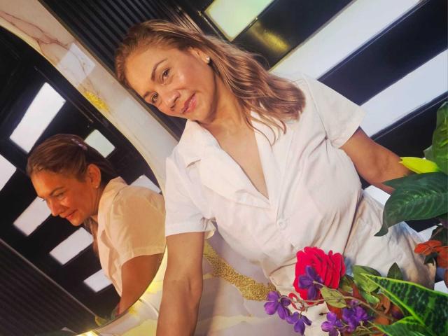 MirandaCeleste, com cabelo loiro, blusa branca e sorriso discreto, segurando flores coloridas, refletida em um espelho. Seios visíveis no reflexo, olhando para baixo, com folhas verdes em primeiro plano..