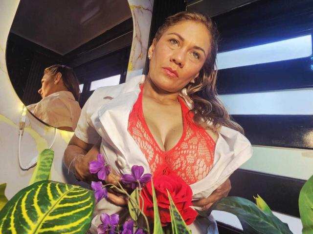 MirandaCeleste, con la pelle abbronzata, reggiseno di pizzo rosso che mette in mostra il suo seno prosperoso, camicia bianca aperta, che tiene fiori viola, che guarda la fotocamera, riflesso nello specchio che mostra la sua schiena, persiane nere..