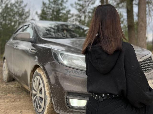 Teresa Ghamdi, met bruin haar dat tot op haar schouders reikt, staat voor een viepe, grijze SUV. Ze draagt een zwarte hoodie en een riem met metalen studs en kijkt naar de voorkant van de auto..