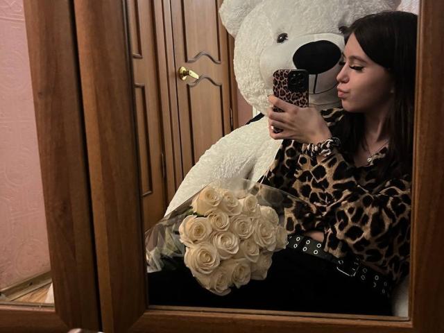 Teresa Ghamdi, avec ses cheveux bruns, un haut à imprimé léopard, une ceinture noire, tenant des roses blanches, se prend en selfie devant un miroir, avec un grand ours en peluche blanc en arrière-plan, une porte en bois et un mur rose..