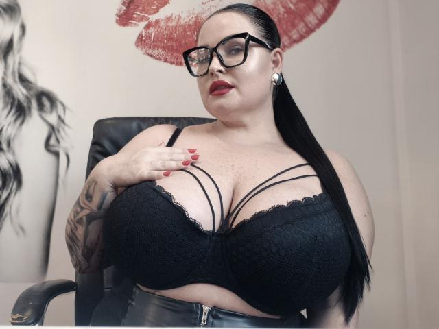 GeorgiaLerox has huge tits in a black lace bra, wearing glasses, red lipstick, and black leather pants, with tattooed arms, long black hair in a ponytail, and red nails.