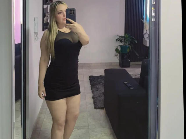 SamyLm draagt een zwarte jurk met een doorschijnende bovenkant, waarbij haar grote billen en kleine vagina zichtbaar zijn in een spiegelzelfportret. Ze heeft blond haar, een lichte huid, houdt een telefoon vast en staat in een kamer met een bank..
