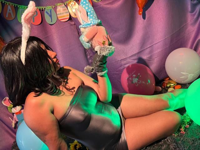 ZarayWhitMoreLove in a gray, shiny, tight bodysuit with bunny ears, holding pink and blue stuffed animals, sitting with green and purple lighting, showing her ass.
