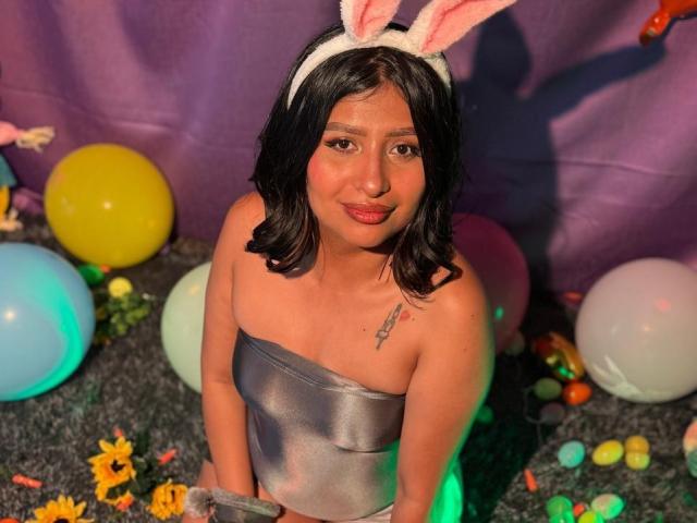 ZarayWhitMoreLove, wearing a silver strapless dress, white bunny ears, and holding a gray furry tail, with a small tattoo on the right shoulder. Colorful balloons and flowers.