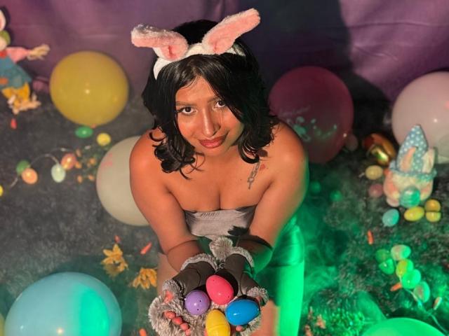 ZarayWhitMoreLove with black curly hair, pink bunny ears, strapless grey dress, holding colorful eggs, surrounded by balloons, green light, and Easter decorations, looking up.