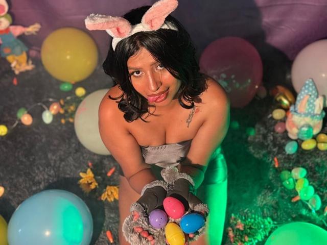 ZarayWhitMoreLove, with black wavy hair, pink bunny ears, and gray strapless outfit, holds colorful Easter eggs, wearing fuzzy gray gloves, looking up, on green-lit carpet.